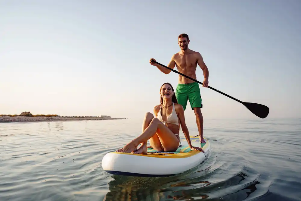 Pár na paddleboarde, muž vesluje a žena sedí, vhodné pre spoločné dobrodružstvo.